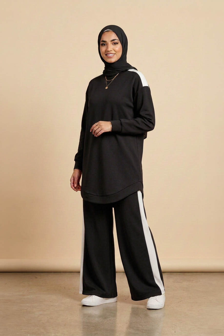 A woman modeling a black long-sleeve tunic top with white shoulder stripes, matching black wide-leg pants with white side stripes, and a black hijab, standing against a beige studio backdrop.