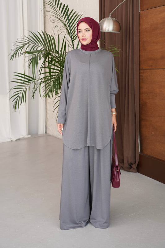 A woman modeling a two-piece gray modest outfit with a high-neck long-sleeve top and wide-leg pants, paired with a deep burgundy hijab and holding a matching handbag in a modern indoor setting.