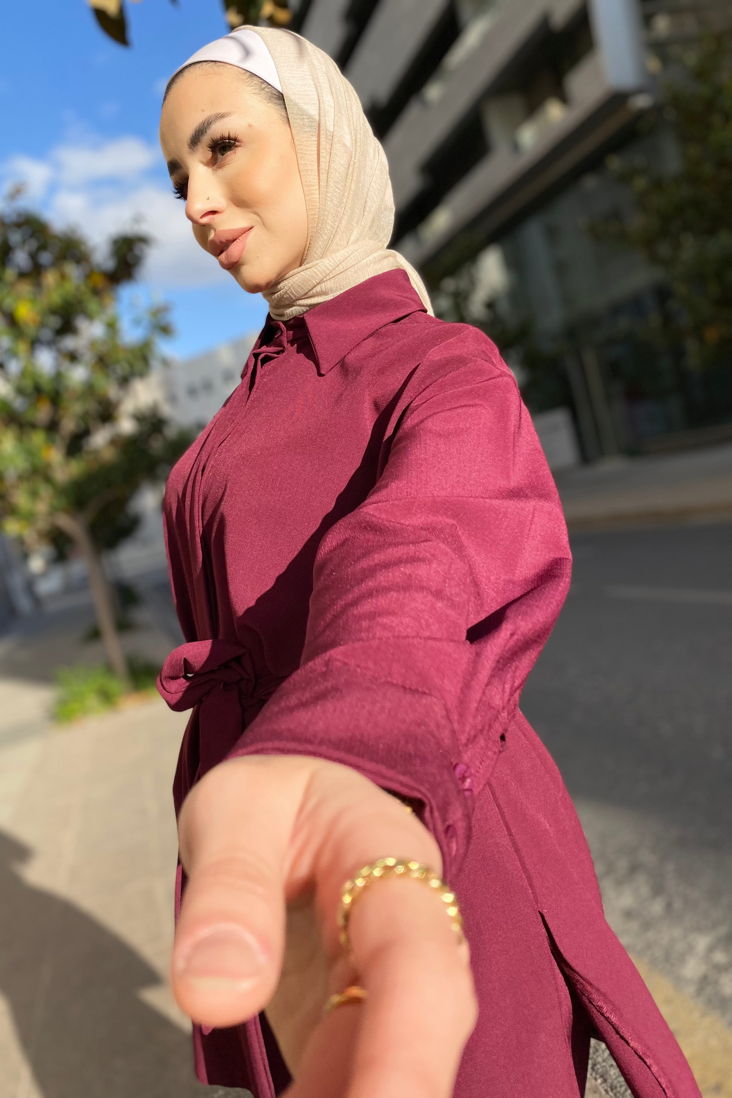 Woman in a burgundy dress and beige hijab taking a selfie outdoors.