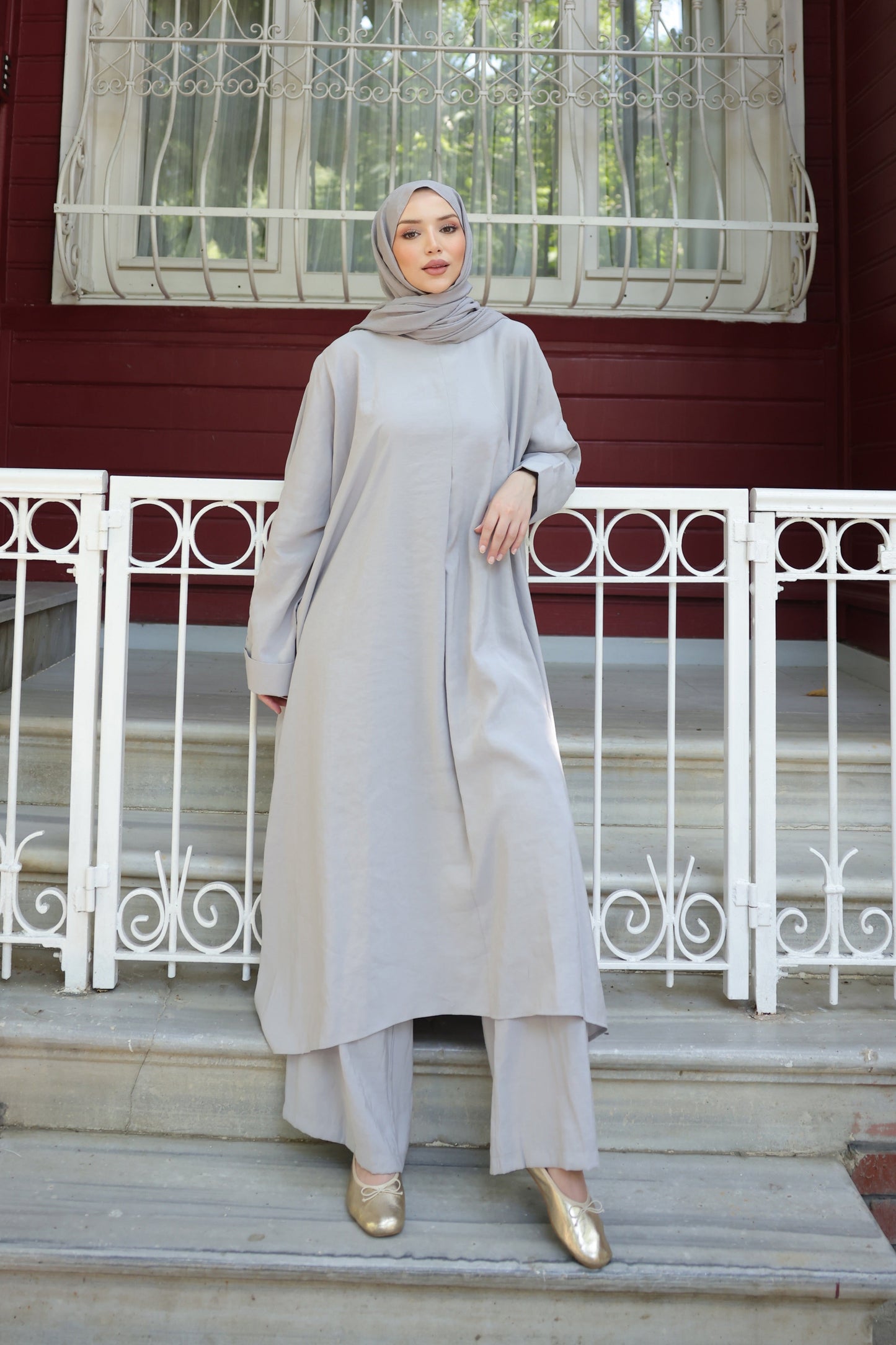 Woman in a light gray outfit standing on steps in front of a red building with white windows.