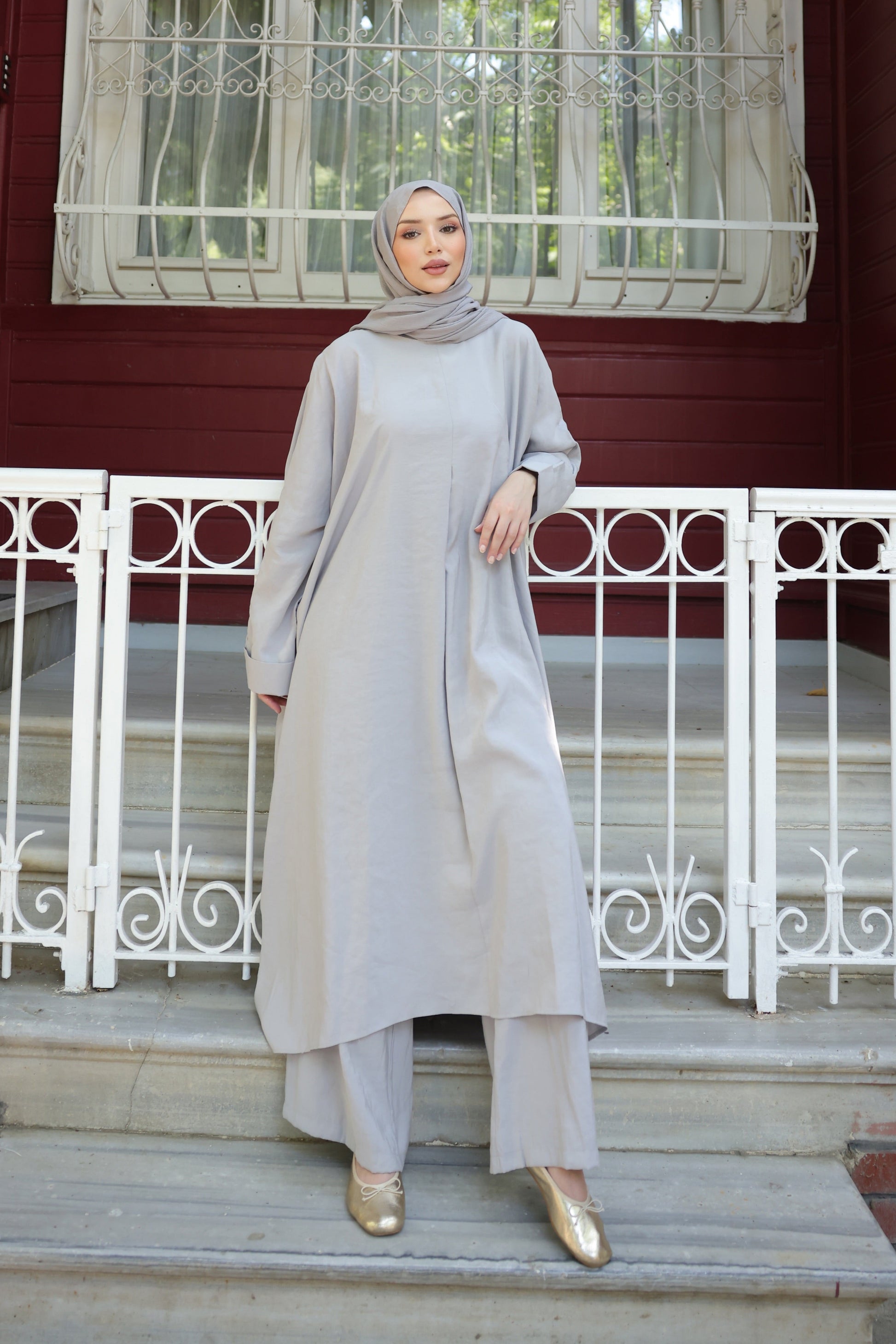 Woman in a light gray outfit standing on steps in front of a red building with white windows.