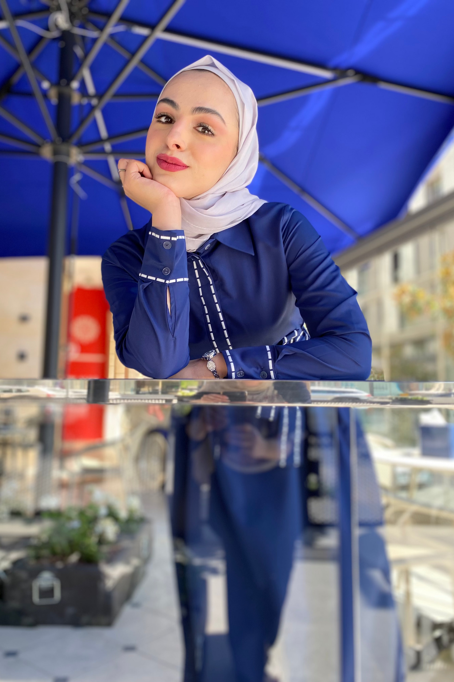 Woman in a blue outfit with a hijab standing under a blue umbrella.