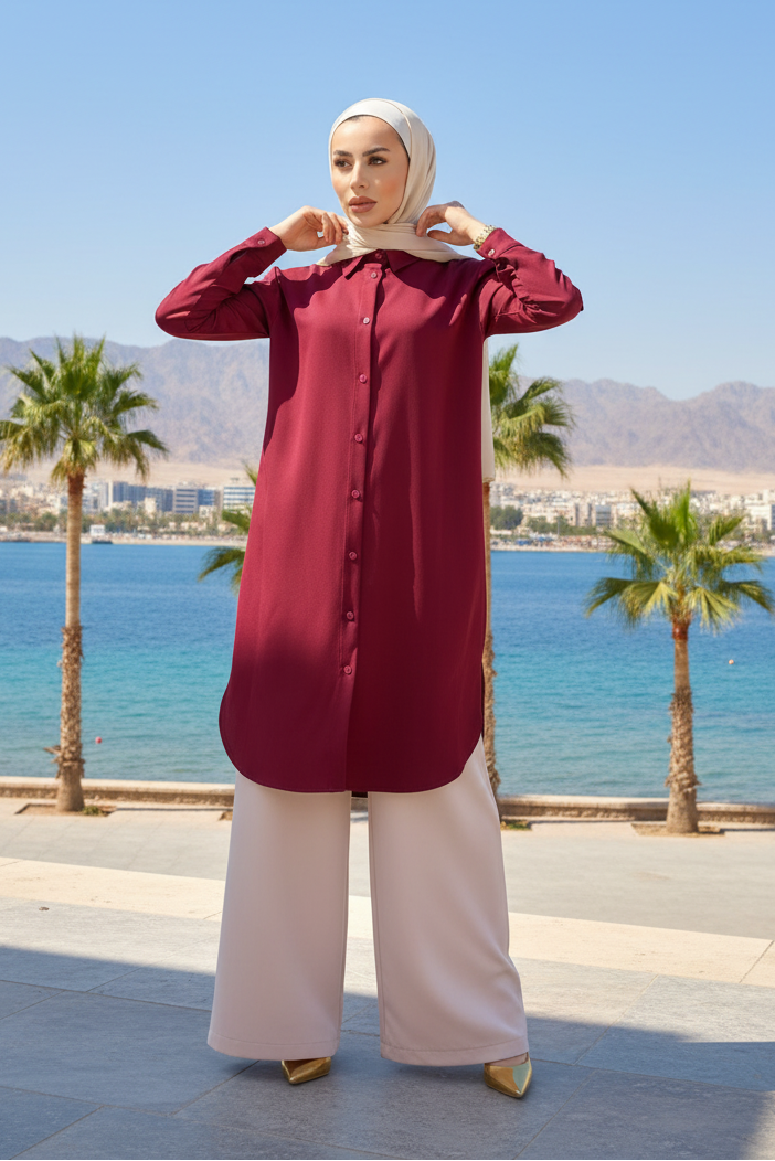 A woman wearing a long-sleeved burgundy button-up tunic and light pink wide-leg pants, paired with a beige hijab, standing on a sunny seaside promenade with palm trees, blue water, and mountains in the background.