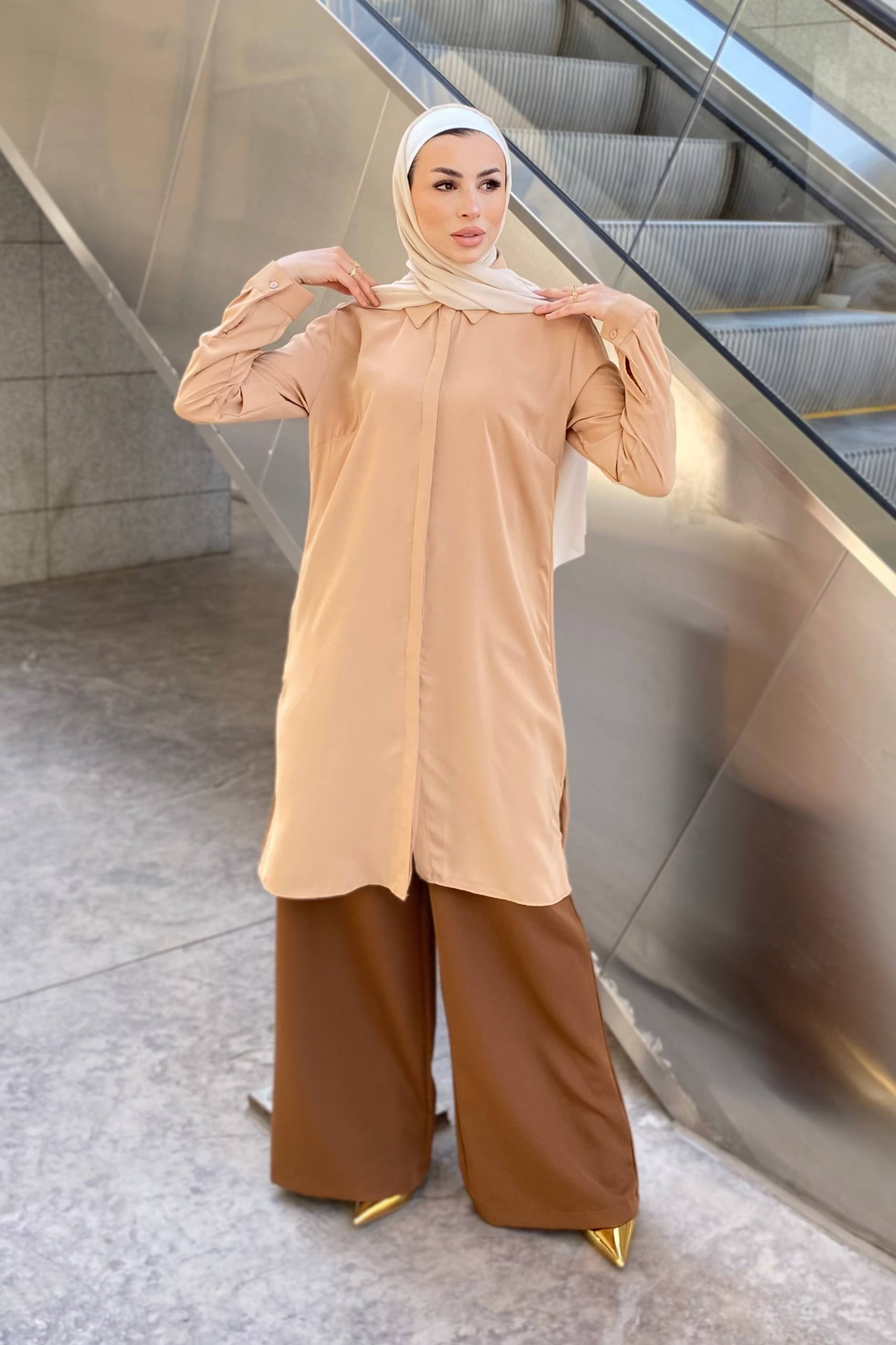 Person wearing a beige top and brown pants standing in front of an escalator.