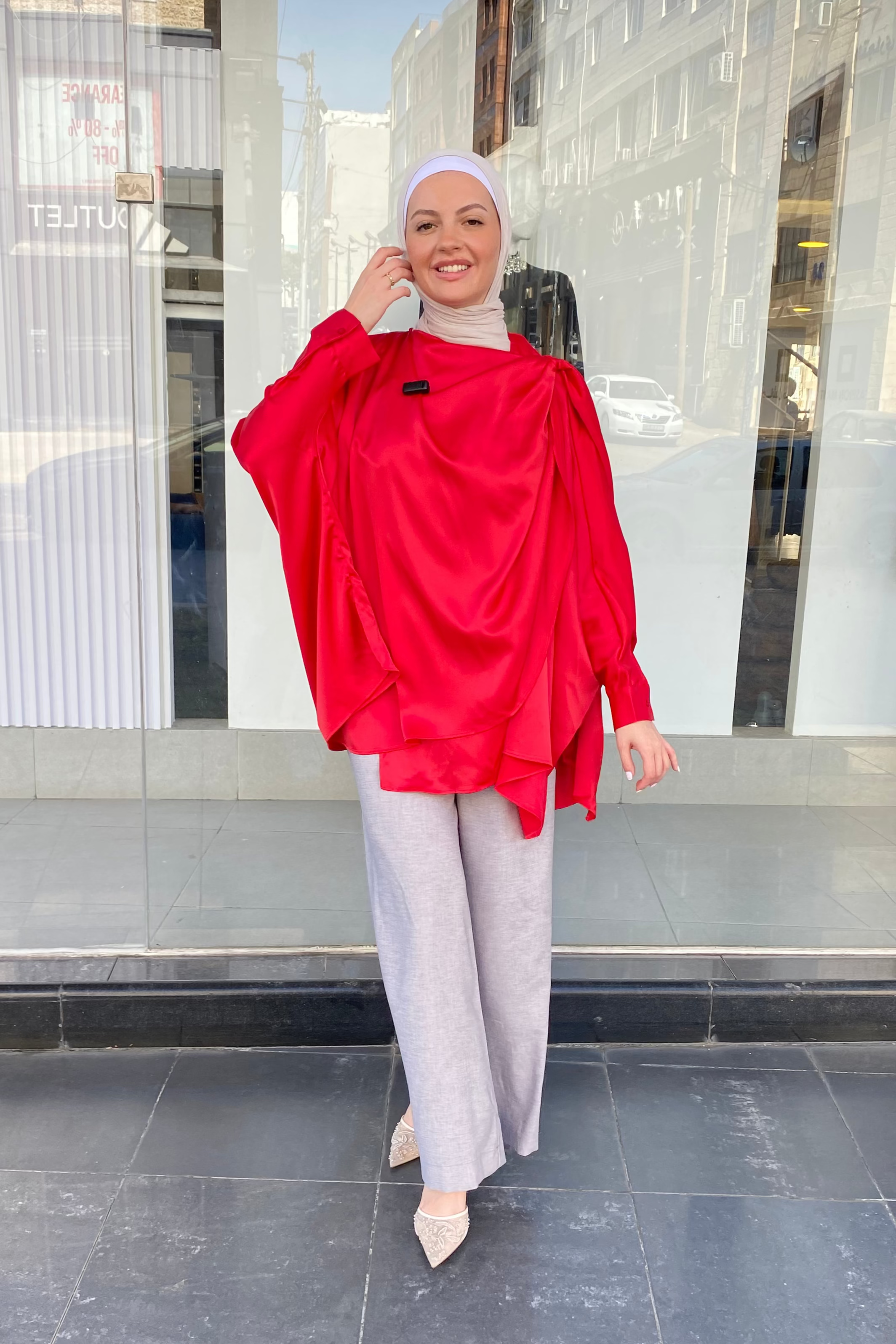 Woman wearing a red top and gray pants standing in front of a store window.