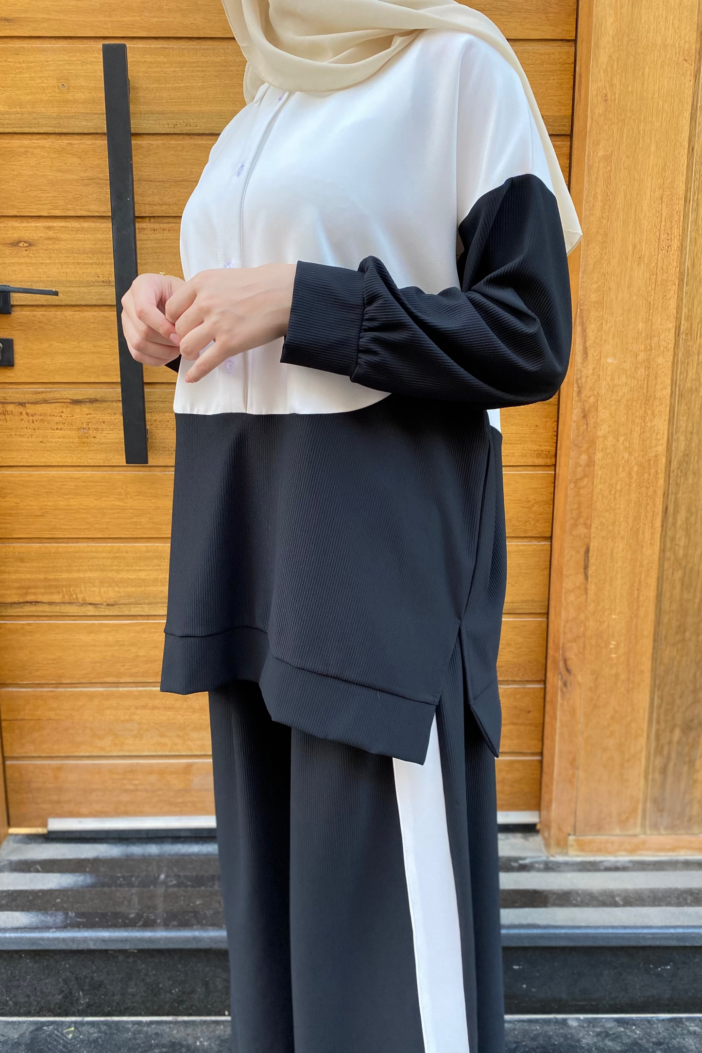 Person wearing a black and white outfit with a hijab in front of a wooden door.