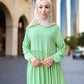 Woman wearing a light green dress and beige hijab standing indoors.