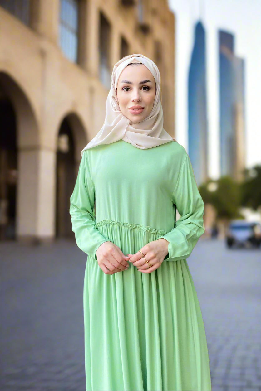Woman wearing a light green dress and beige hijab standing indoors.