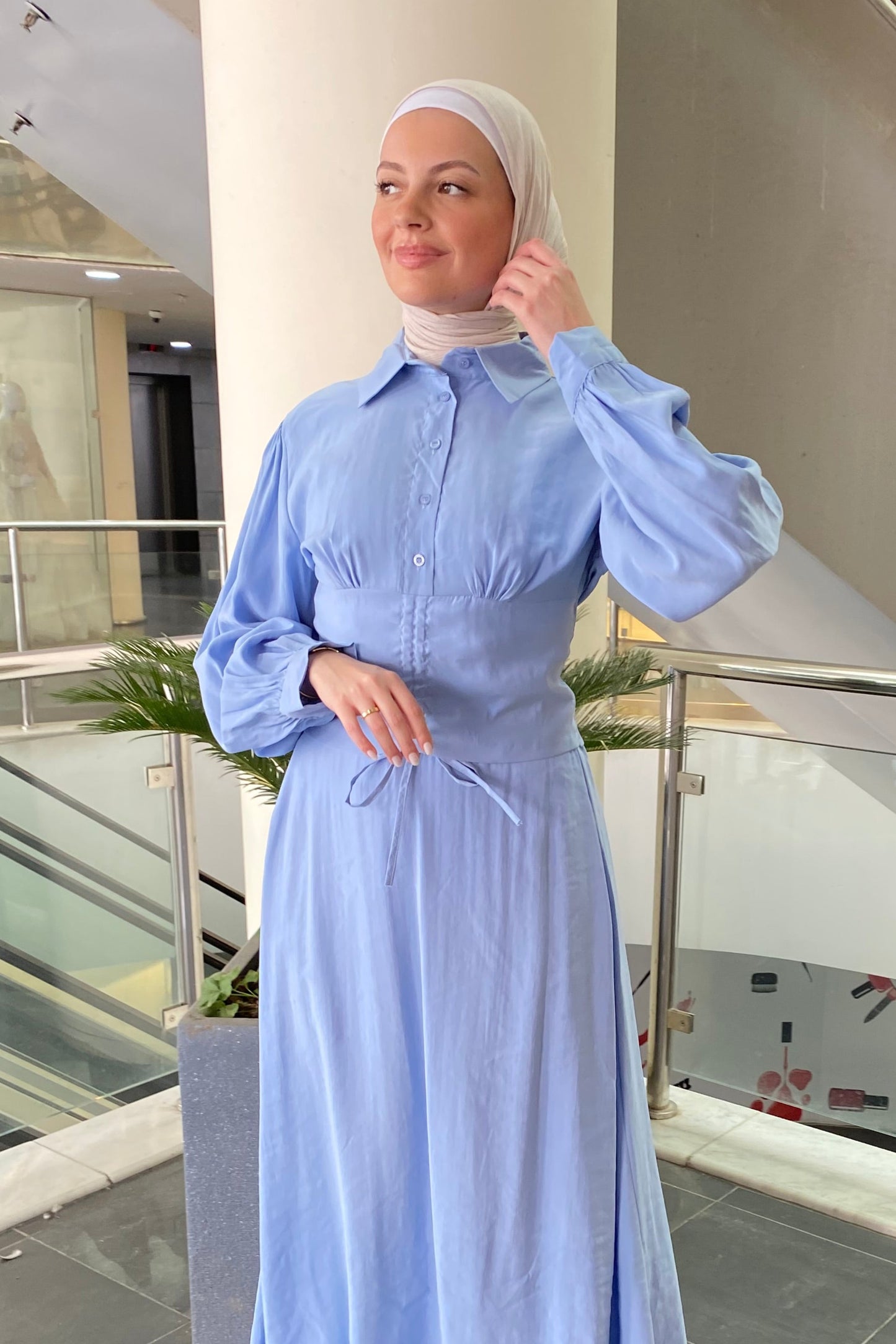 Woman in a light blue dress standing in a modern indoor setting with glass walls.