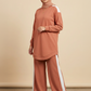 A woman modeling a matching terracotta rust-colored activewear set consisting of a long-sleeve mock-neck tunic with white shoulder panels and matching wide-leg pants with white side stripes, paired with a matching hijab and white sneakers. 