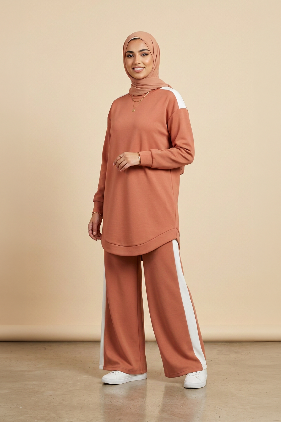 A woman modeling a matching terracotta rust-colored activewear set consisting of a long-sleeve mock-neck tunic with white shoulder panels and matching wide-leg pants with white side stripes, paired with a matching hijab and white sneakers. 