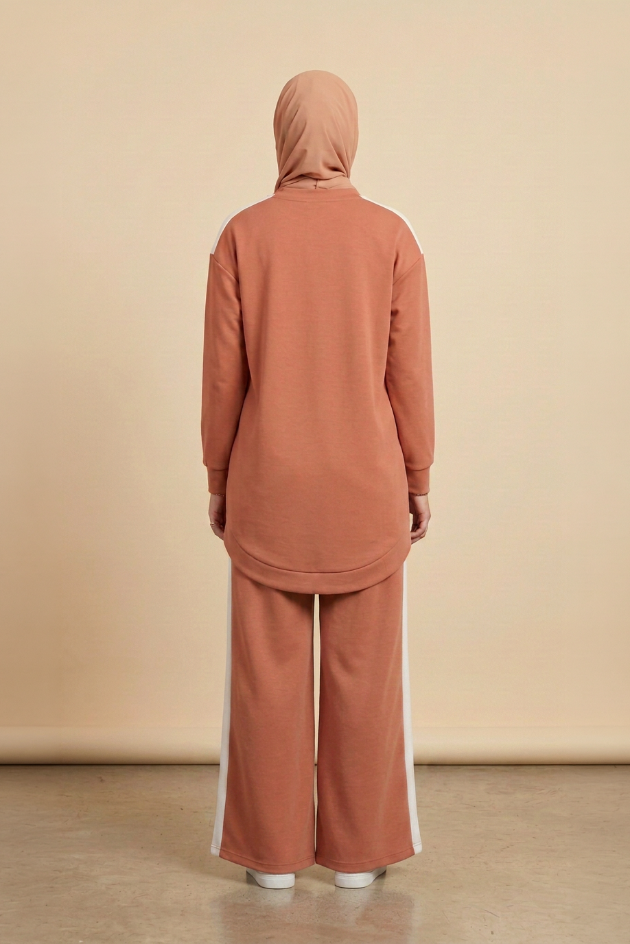 A person viewed from the back wearing a matching terracotta-colored hijab, long-sleeve top, and wide-leg pants with white side stripes, standing against a plain beige studio backdrop.