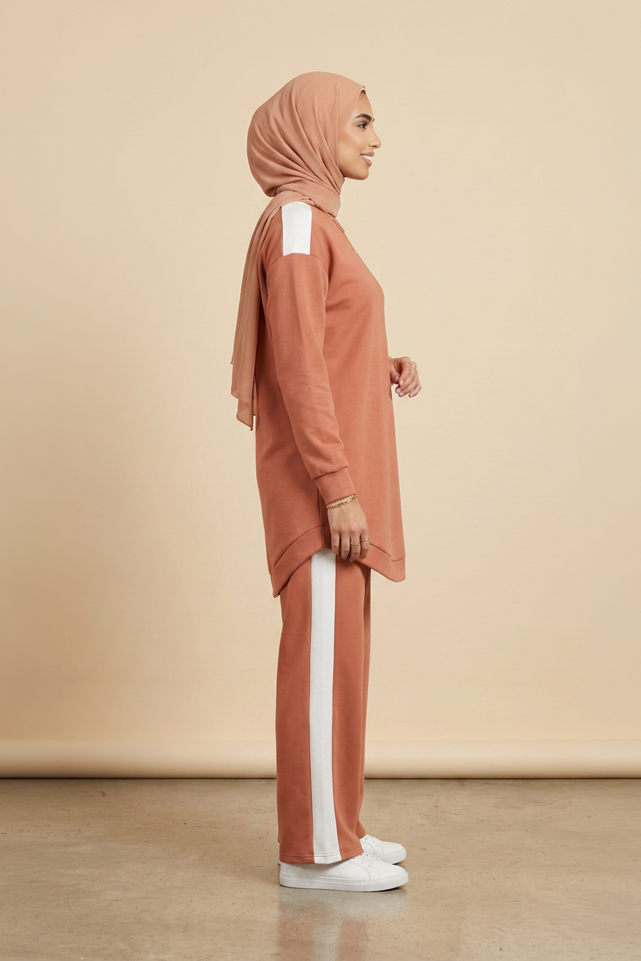 A woman in profile wearing a dusty rose hijab, matching long-sleeve top with white shoulder panels, and matching wide-leg pants with white side stripes, standing against a plain beige studio backdrop.