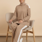 A woman wearing a matching beige hijab, long-sleeve top, and wide-leg pants set with white side stripes, seated on a modern gray chair against a warm beige studio background.