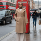Woman in a beige coat standing on a city street with a red phone booth and double-decker bus in the background.