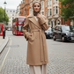 Woman in a beige coat standing on a city street with double-decker buses in the background.