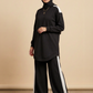 A woman modeling a black long-sleeve tunic top with white shoulder stripes, matching black wide-leg pants with white side stripes, and a black hijab, standing against a beige studio backdrop.