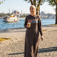 Woman in a long dress standing by a waterfront with trees and a ferry in the background.