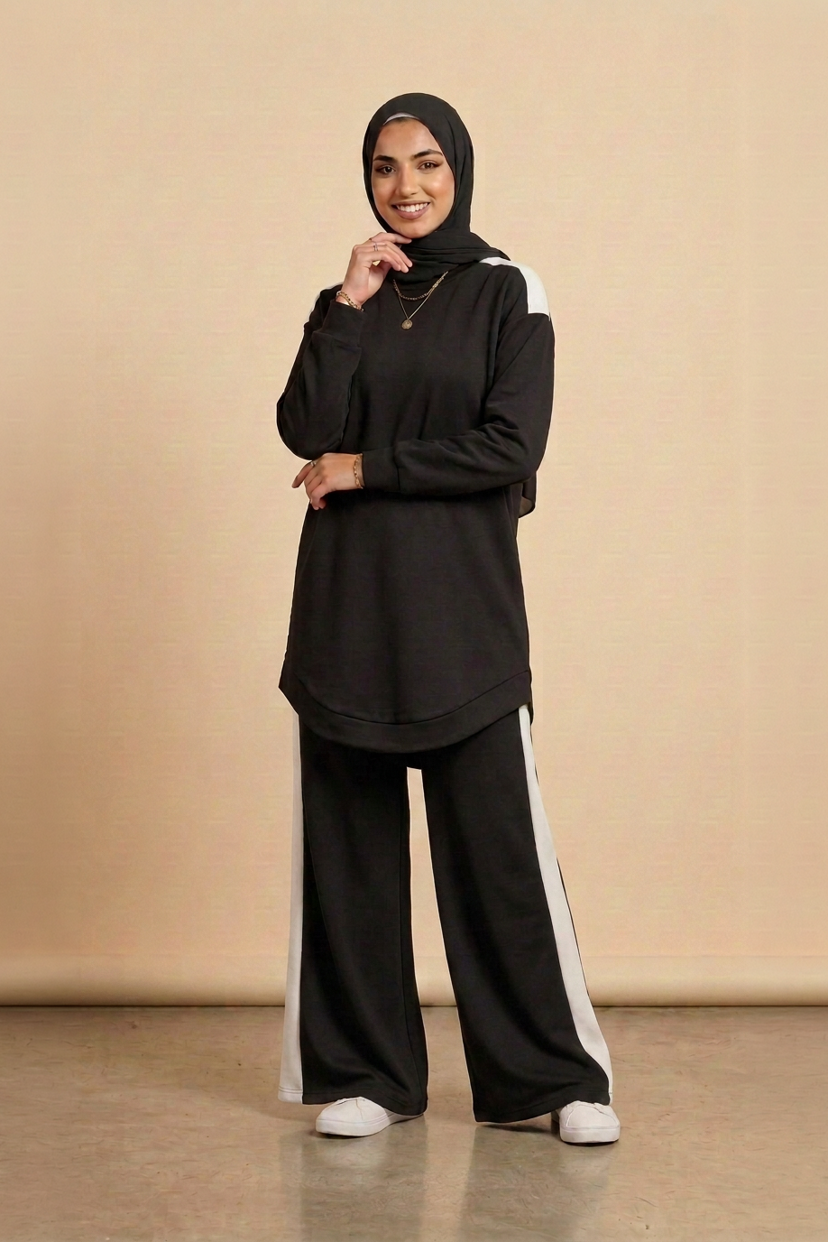 A woman wearing a black long-sleeve tunic with white shoulder panels, matching black wide-leg pants with white side stripes, and a black hijab, standing against a beige studio backdrop.