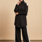 A woman wearing a black long-sleeve tunic with white shoulder panels, matching black wide-leg pants with white side stripes, and a black hijab, standing against a beige studio backdrop.