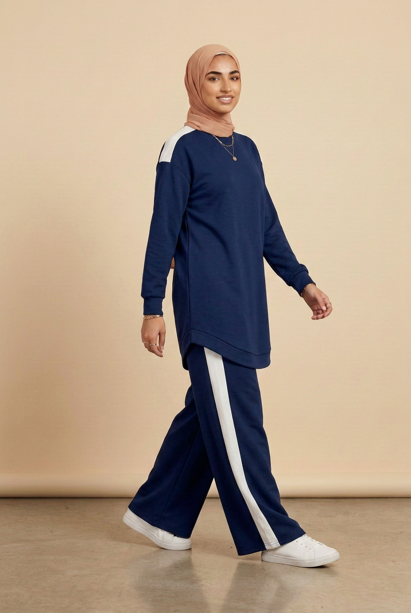 A woman wearing a navy blue long-sleeve tunic top with white shoulder stripes and matching navy wide-leg pants with white side stripes, paired with a beige hijab and white sneakers, walking against a solid beige studio background.