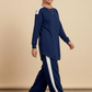 A woman wearing a navy blue long-sleeve tunic top with white shoulder stripes and matching navy wide-leg pants with white side stripes, paired with a beige hijab and white sneakers, walking against a solid beige studio background.