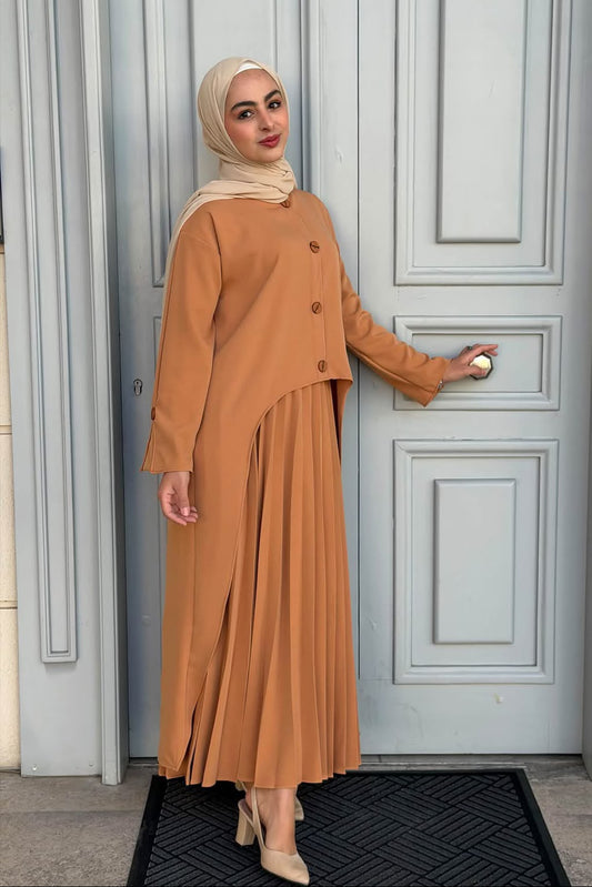A woman wearing a tan long-sleeve button-front abaya dress with a matching pleated skirt and beige hijab stands in front of a light gray paneled door, holding the doorknob. 