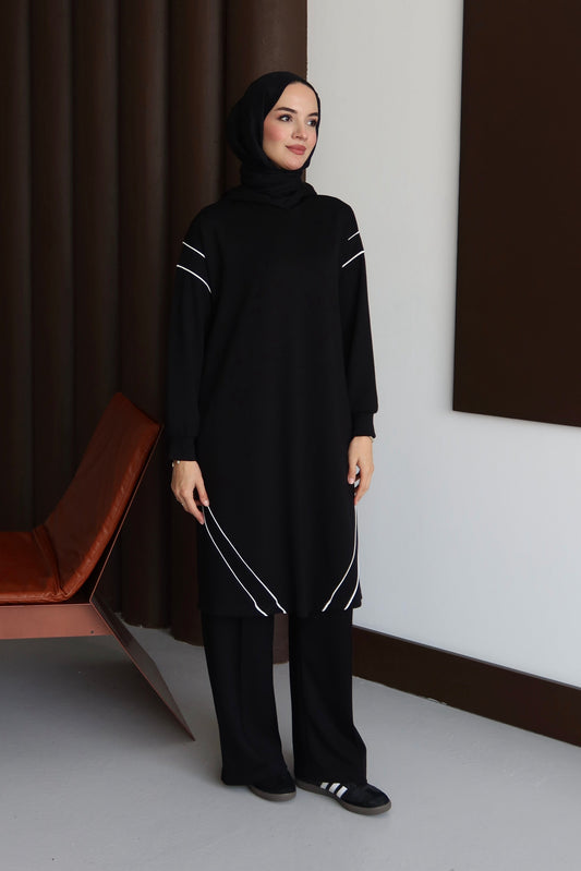 Woman in a black outfit standing in a minimalistic room with a brown bench and white wall.