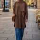A woman wearing a brown long-sleeve tunic with a draped collar, a matching brown hijab, and blue jeans walking on a city street.