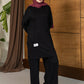 A woman wearing a black long-sleeve tunic and wide-leg pants with a dusty rose hijab and sunglasses, standing in front of a wooden paneled wall.