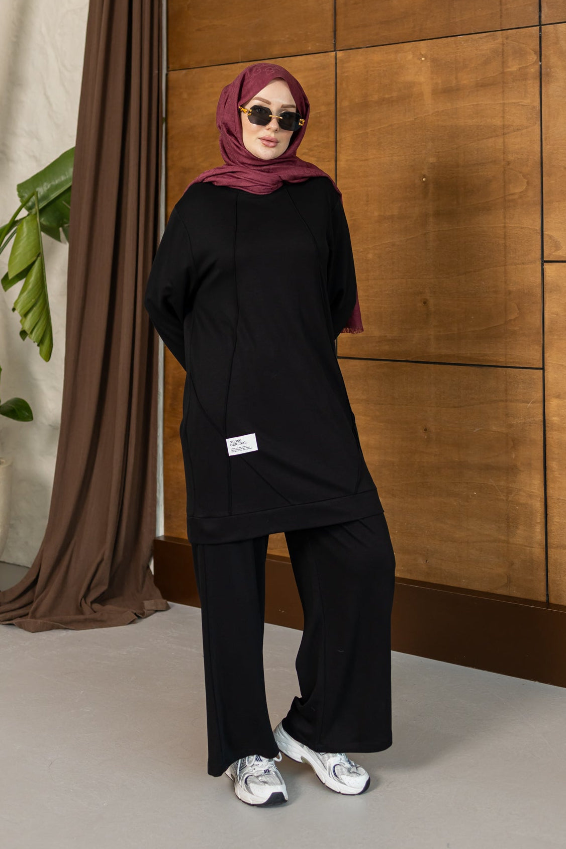 A woman wearing a black long-sleeve tunic and wide-leg pants with a dusty rose hijab and sunglasses, standing in front of a wooden paneled wall.