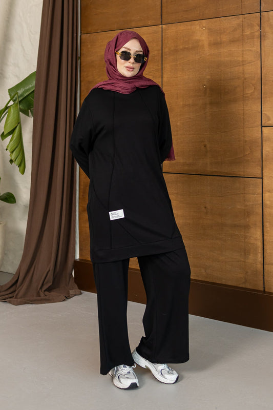 A woman wearing a black long-sleeve tunic and wide-leg pants with a dusty rose hijab and sunglasses, standing in front of a wooden paneled wall.
