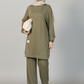 A woman modeling a matching olive green lounge set consisting of a long-sleeve tunic top and wide-leg pants, paired with a light beige hijab and a small cream drawstring bag, against a solid gray studio background.
