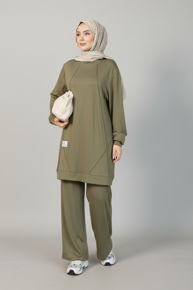 A woman modeling a matching olive green lounge set consisting of a long-sleeve tunic top and wide-leg pants, paired with a light beige hijab and a small cream drawstring bag, against a solid gray studio background.