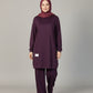 A woman wearing a matching deep purple modest outfit with a coordinating hijab, standing in an indoor setting with a wood-paneled wall, brown curtain, and potted plant.