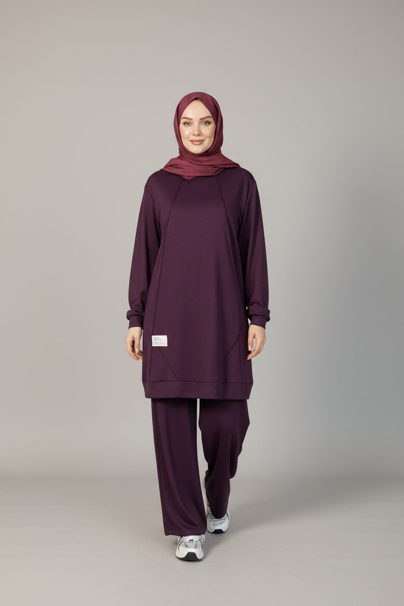 A woman wearing a matching deep purple modest outfit with a coordinating hijab, standing in an indoor setting with a wood-paneled wall, brown curtain, and potted plant.