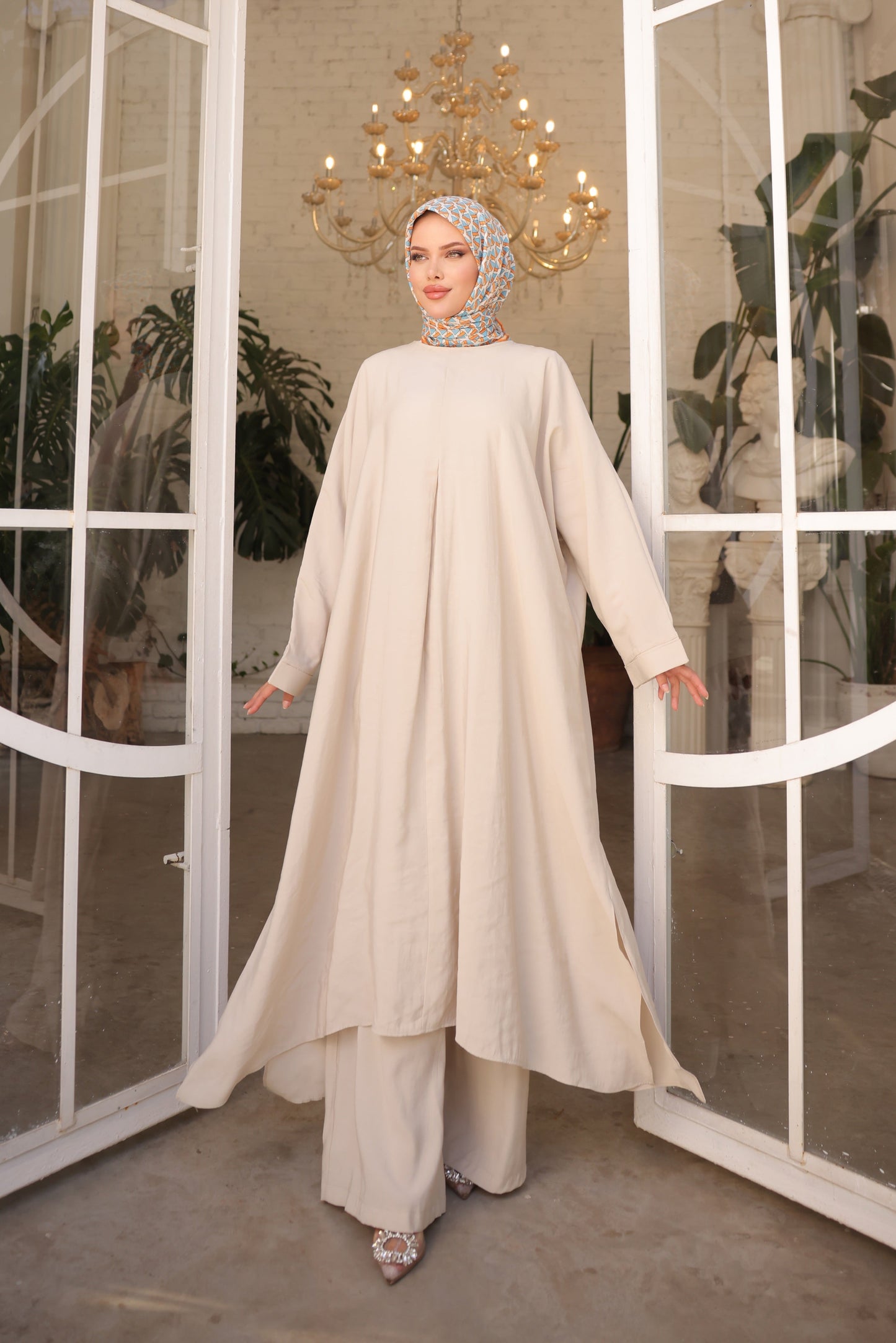Woman in a white outfit standing in front of a large window with a chandelier above.