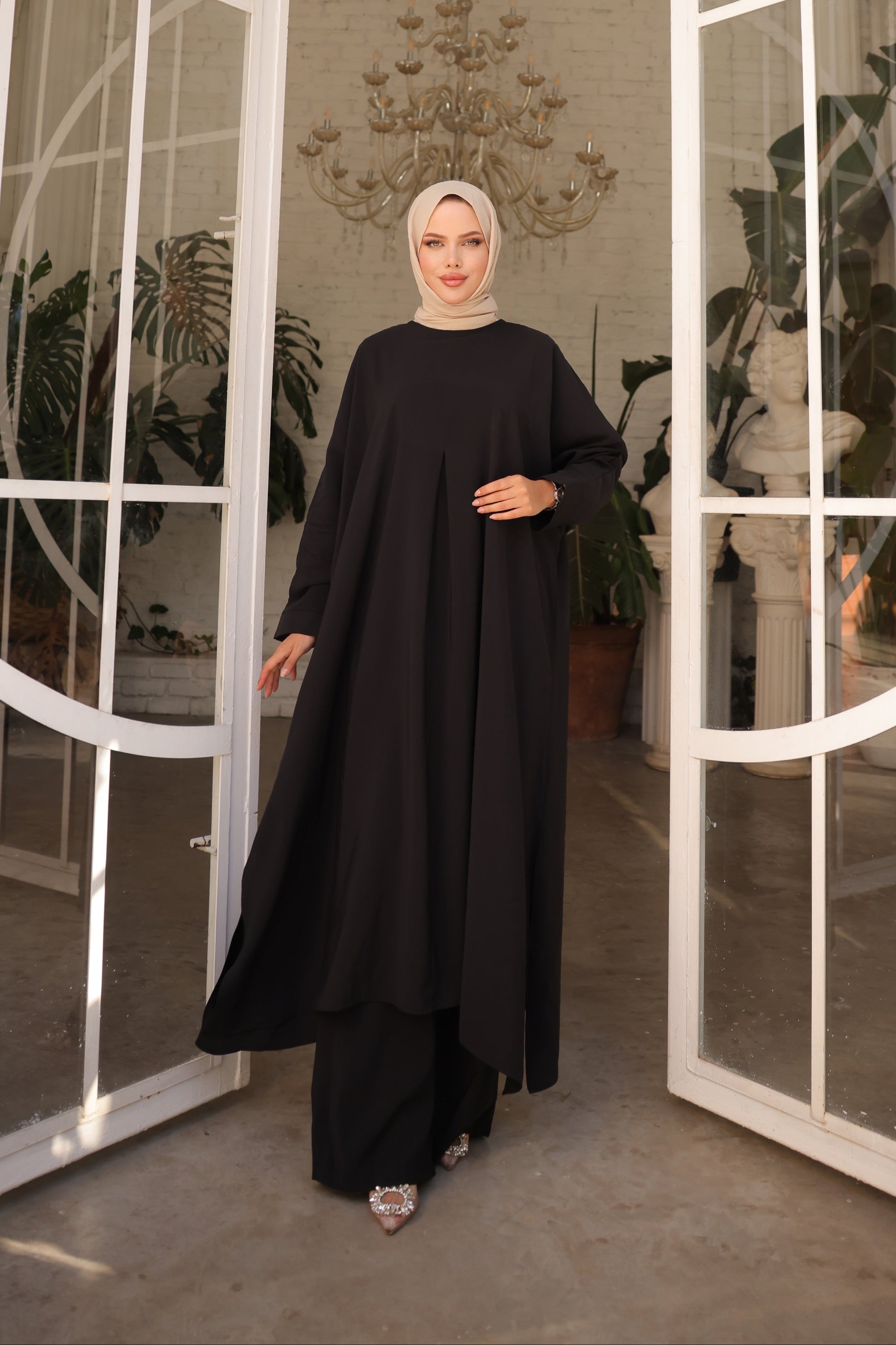 Woman in a black outfit standing in a modern interior setting with large glass doors and plants.